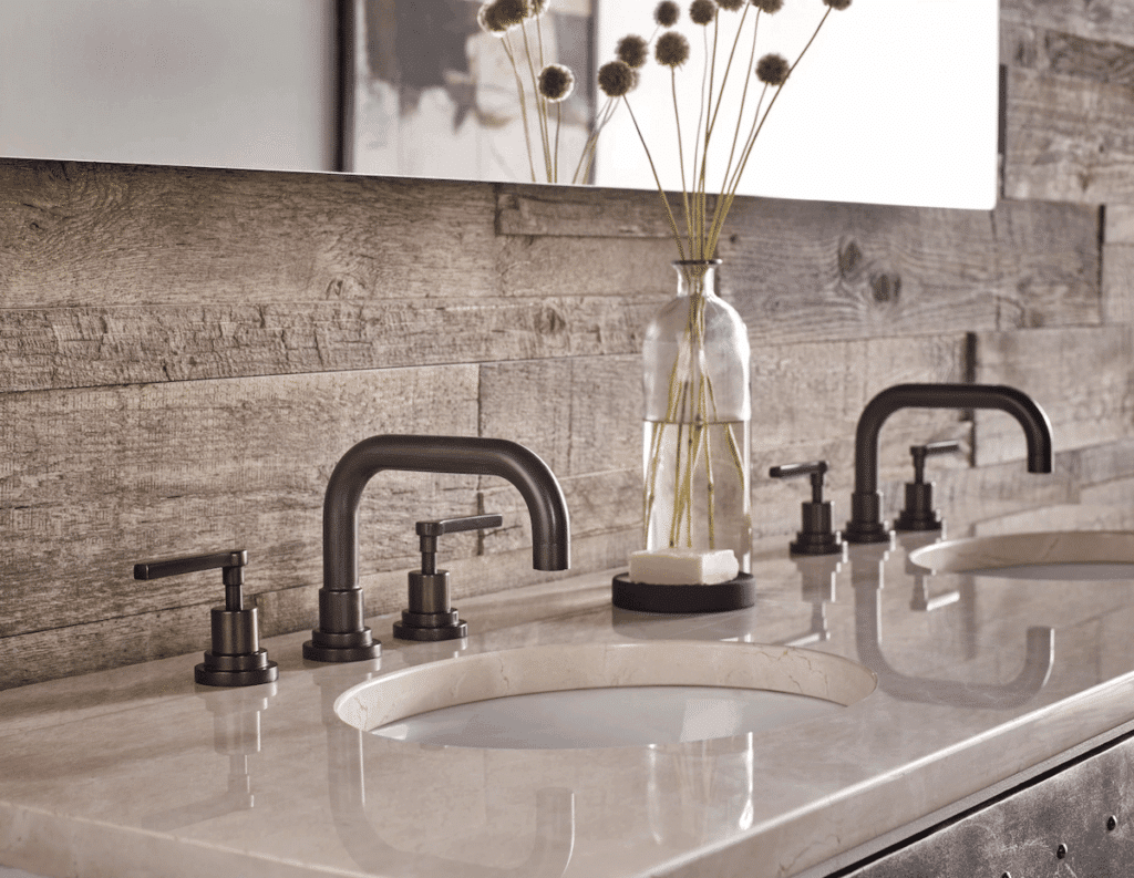 Close-up of dark bronze widespread bathroom faucets on a cream marble vanity with a rustic, reclaimed wood backsplash.