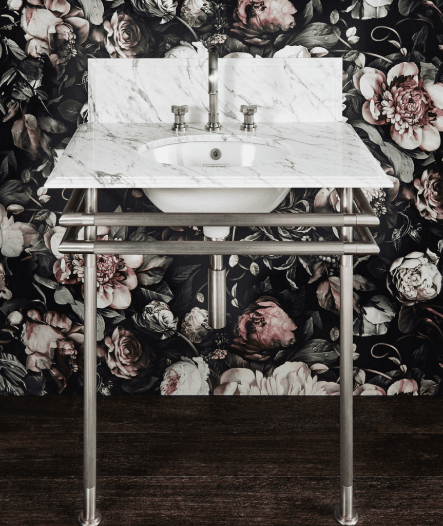 White marble console sink with ribbed silver legs and cross-handle faucet, set against a dark floral wallpaper background.